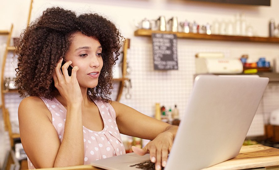 Small business owner in her coffee shop typing on her laptop while talking on the phone